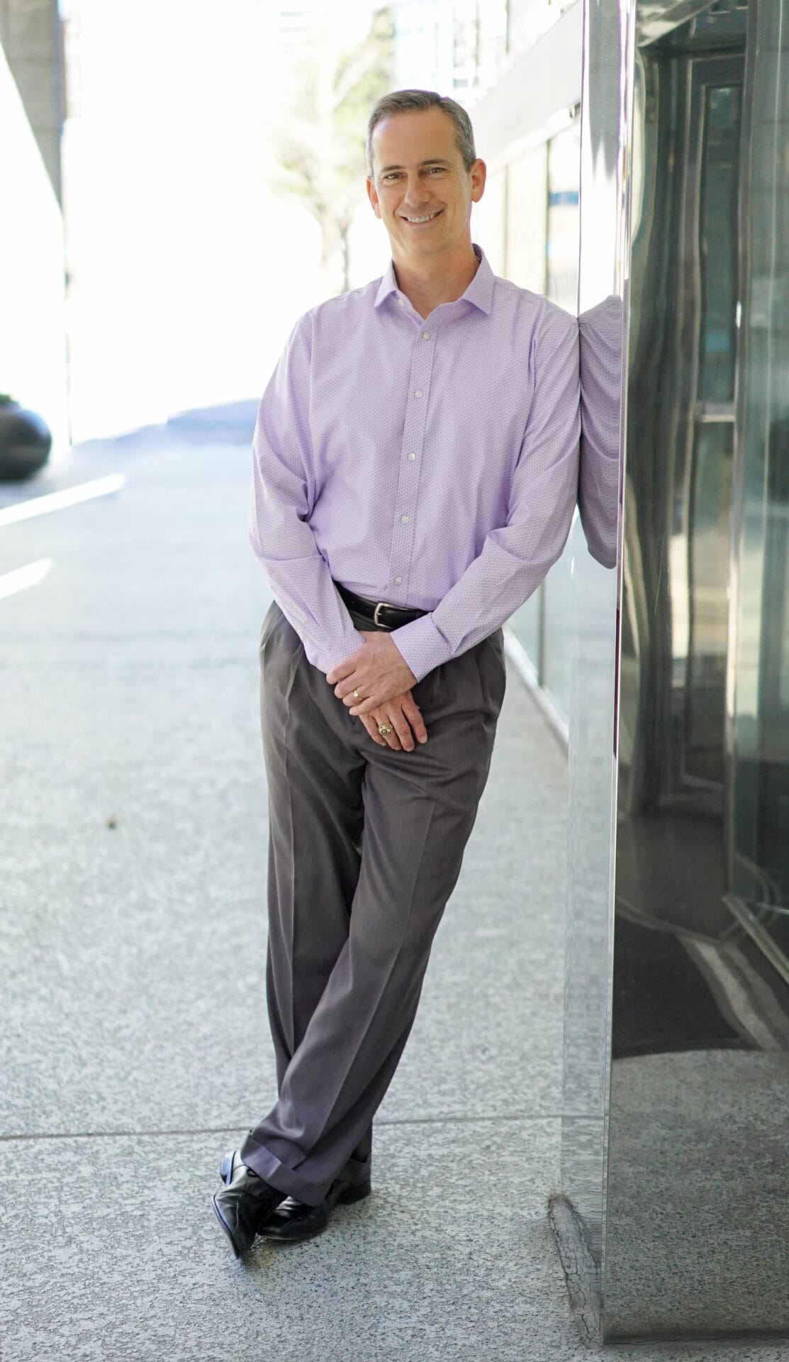 A Caucasian male in a purple polo and gray slacks leaning against a wall smiling for a headshot